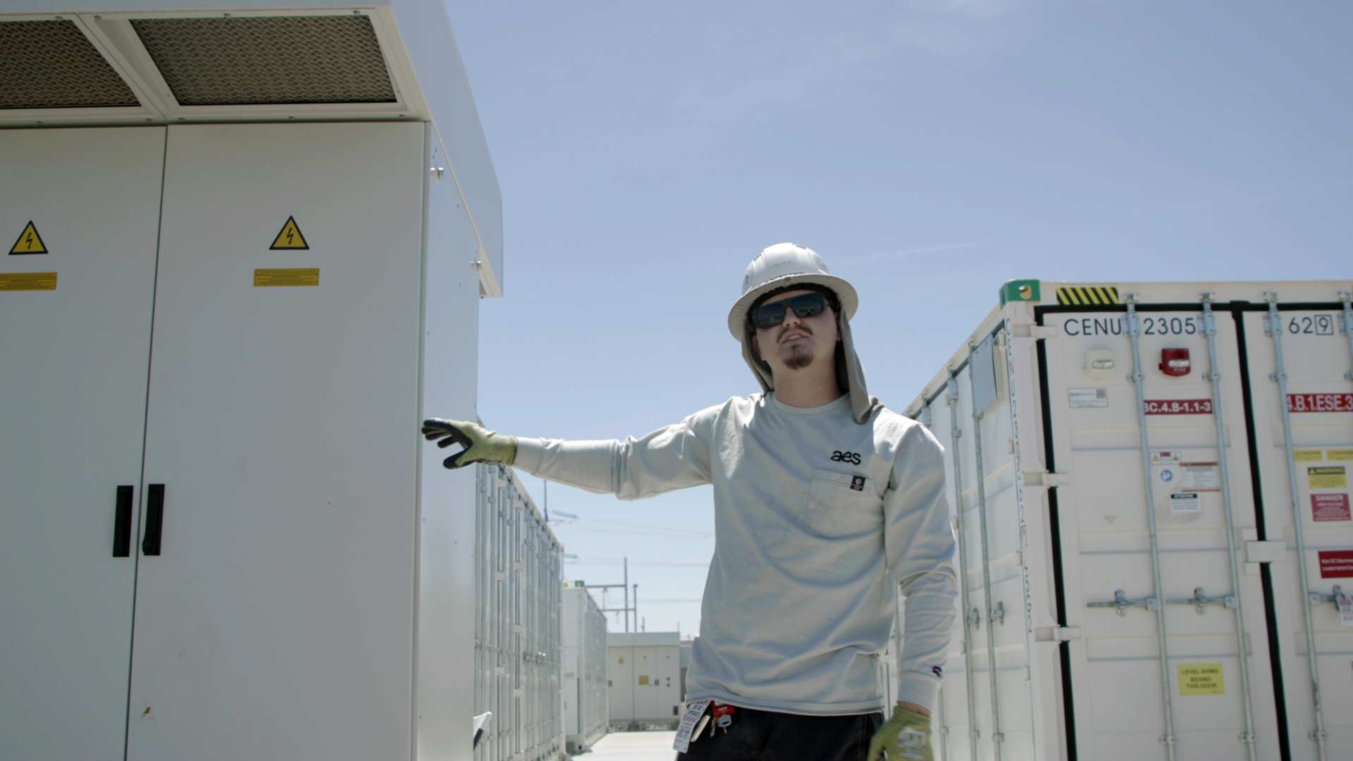 Battery energy storage technician, Tyler, stands in the sun in front of two battery storage units in Arizona.